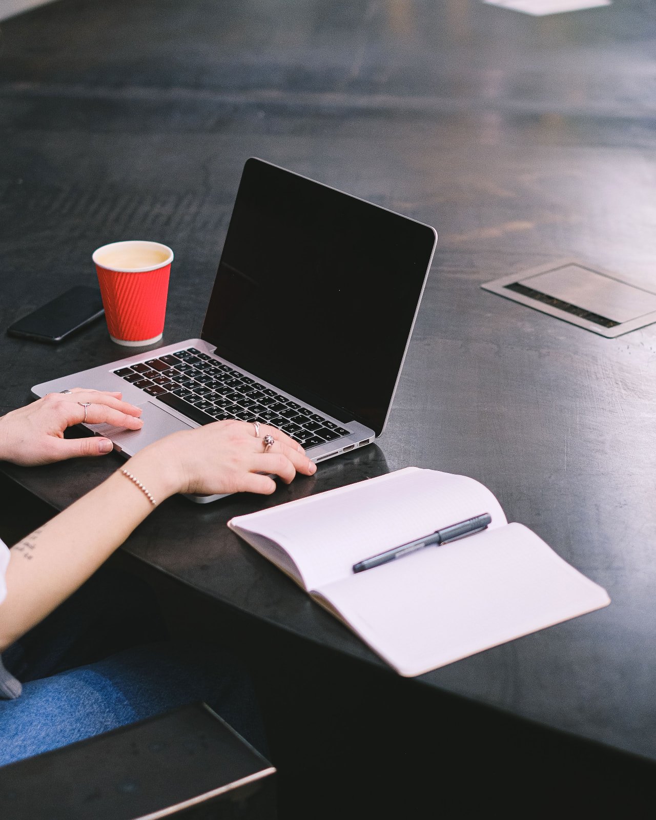 A laptop, open notebook, phone and coffee cup on a dark desk as someone works